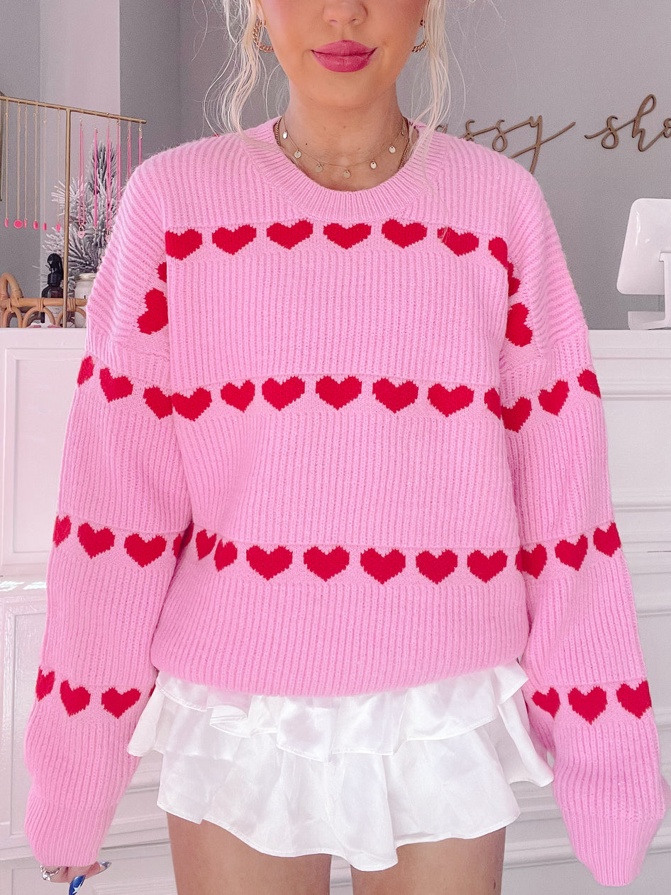 Pink knit sweater with red heart accents; styled with a white ruffled skirt.
