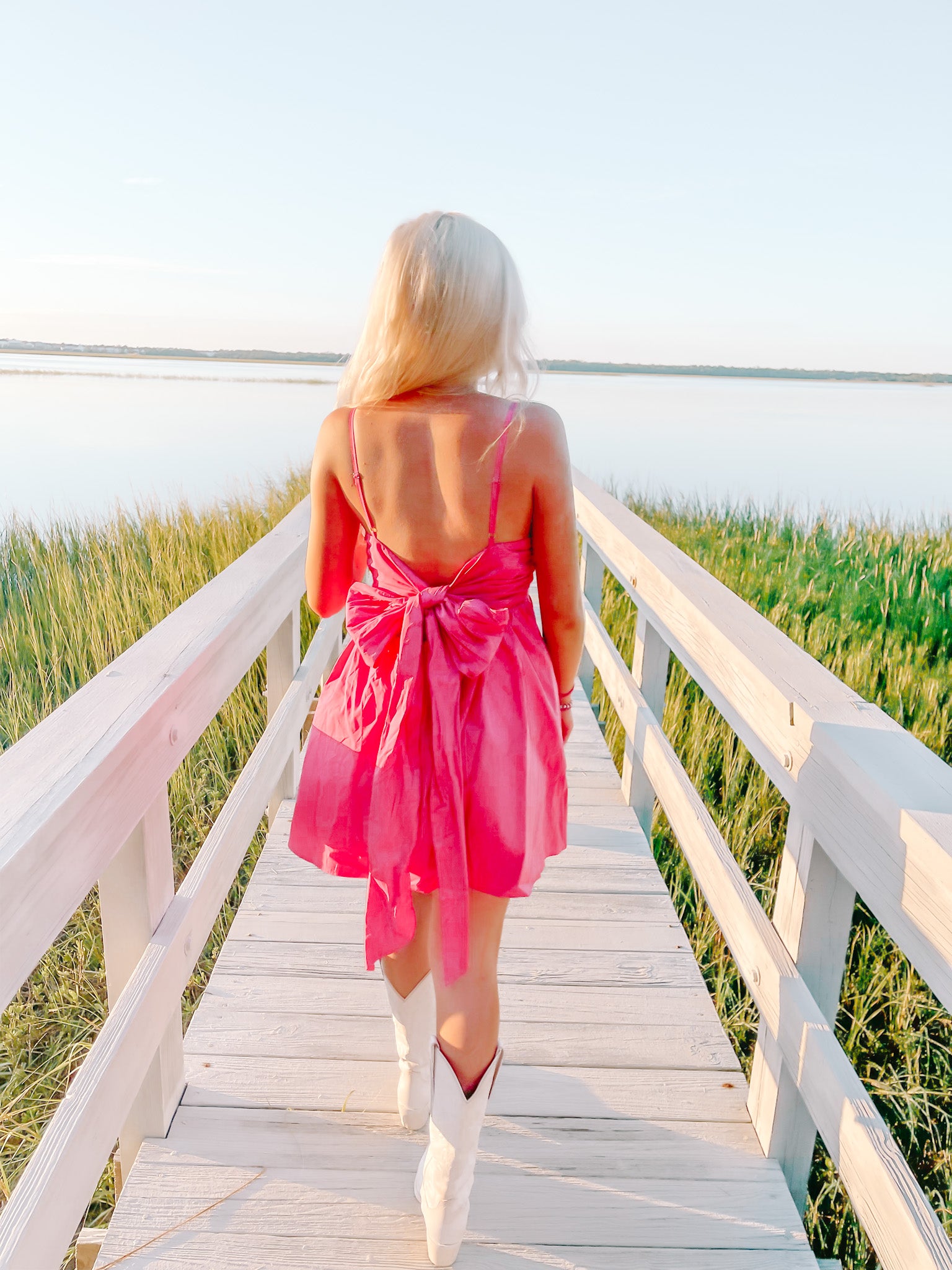 Pink bow-back dress worn on a boardwalk overlooking a tranquil lake at sunset.
