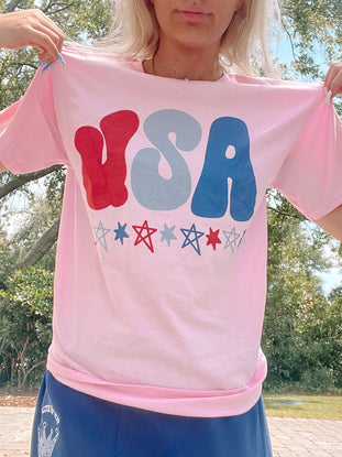 Light pink tee with retro-style USA graphic and stars; outdoors setting.
