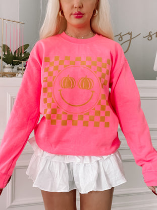 Hot pink crewneck sweatshirt featuring a gold pumpkin smiley face on a checkered background.
