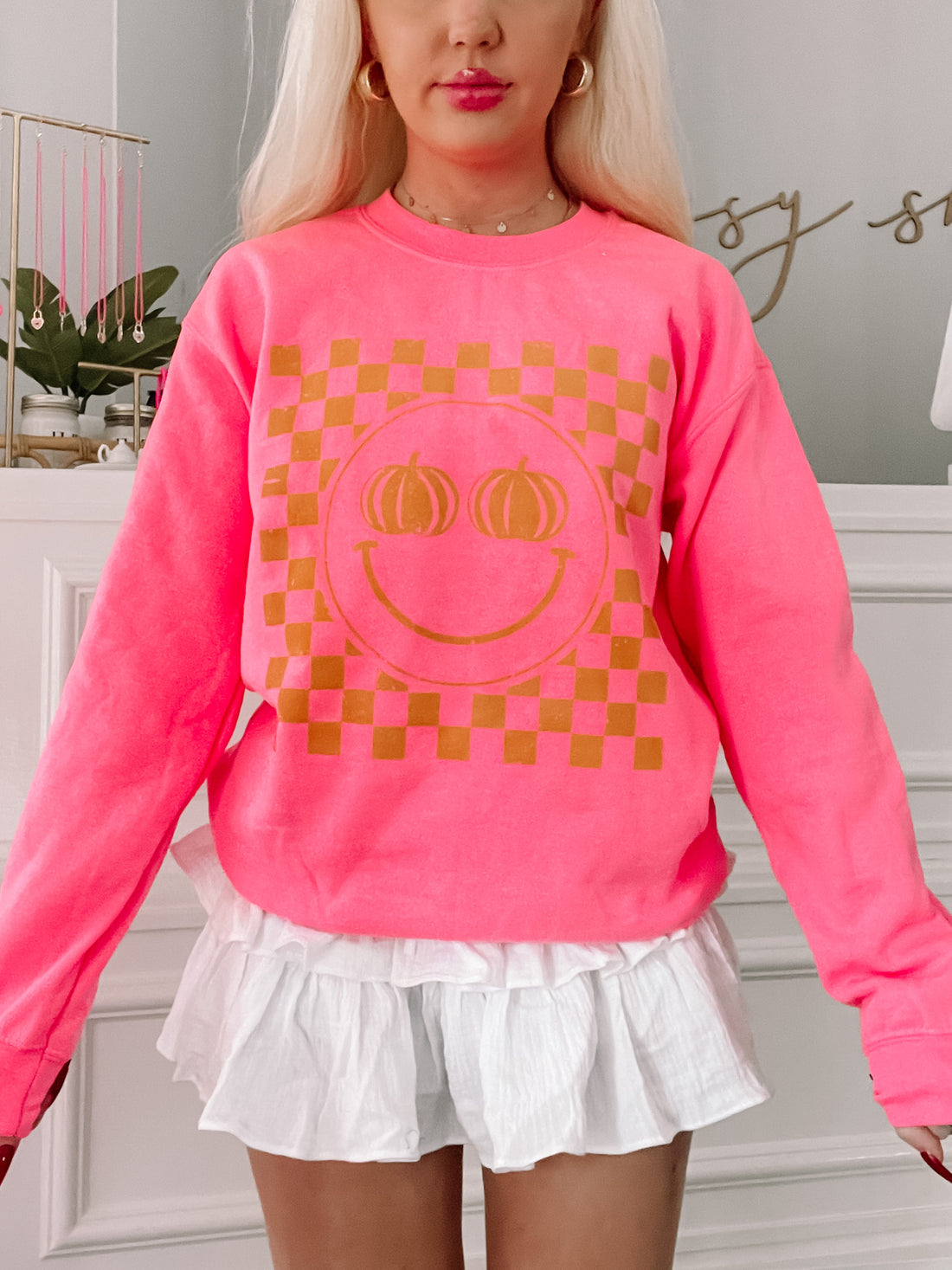 Hot pink crewneck sweatshirt featuring a gold pumpkin smiley face on a checkered background.
