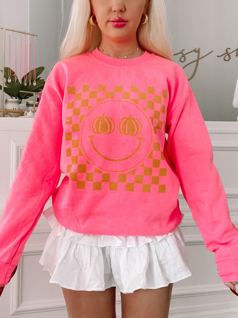 Hot pink crewneck sweatshirt featuring a gold pumpkin smiley face on a checkered background.
