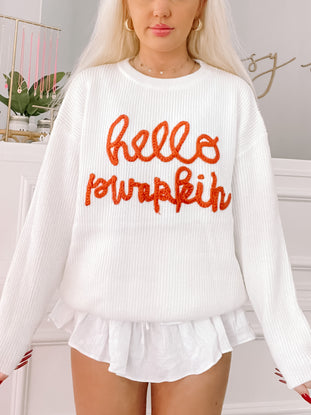 Cream-colored knit sweater featuring an orange "Hello Pumpkin" design; styled with a white ruffle-hemmed undershirt.
