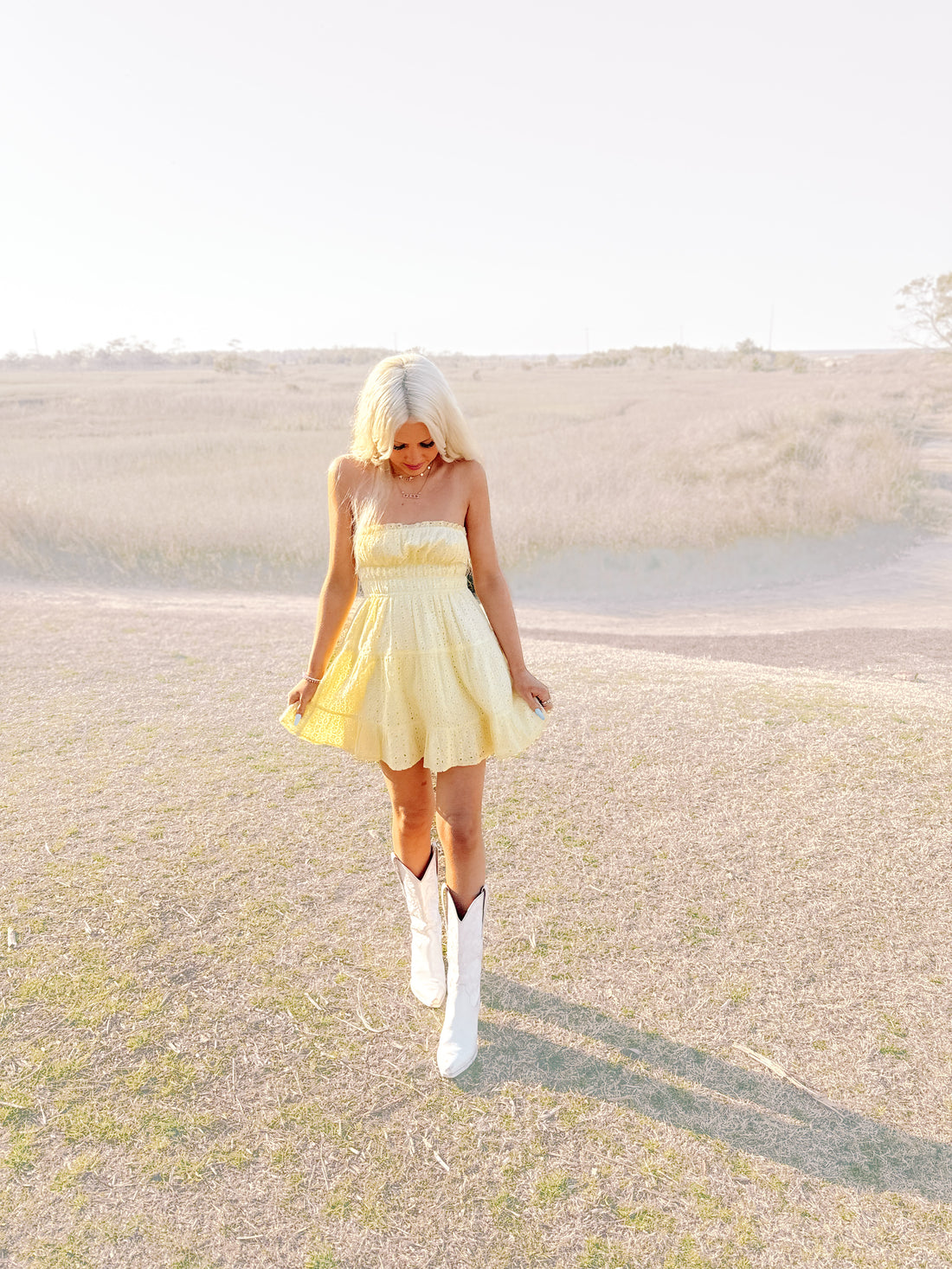 Pale yellow eyelet sundress styled outdoors, paired with white cowboy boots in a grassy field.
