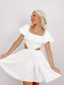 White eyelet mini dress with puff sleeves and cutout detail, modeled against a white background.
