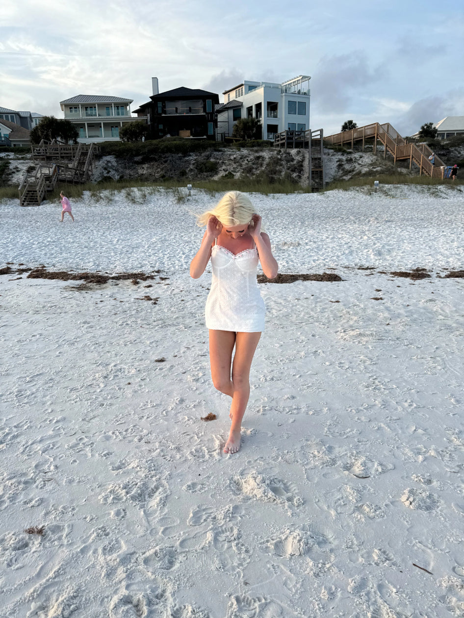 White eyelet mini dress on a beach, near upscale homes.
