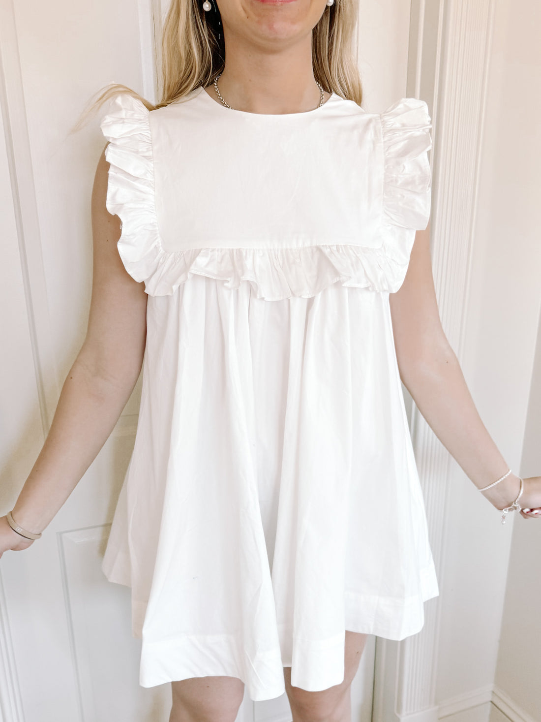 White eyelet mini dress with ruffle details, shown on model against white door.
