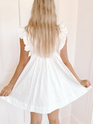 White eyelet mini dress with ruffle sleeves, shown from the back against a white wall.
