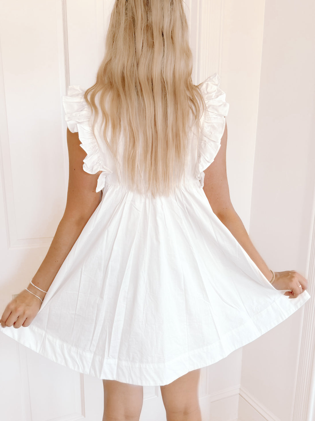 White eyelet mini dress with ruffle sleeves, shown from the back against a white wall.

