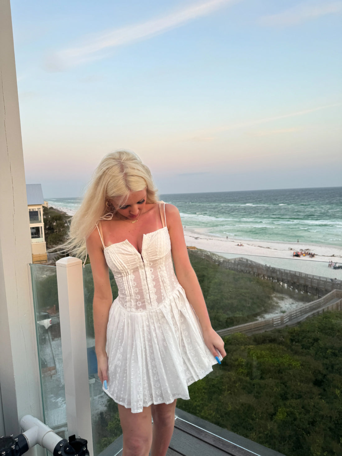 White eyelet mini dress on a beachside balcony at sunset.
