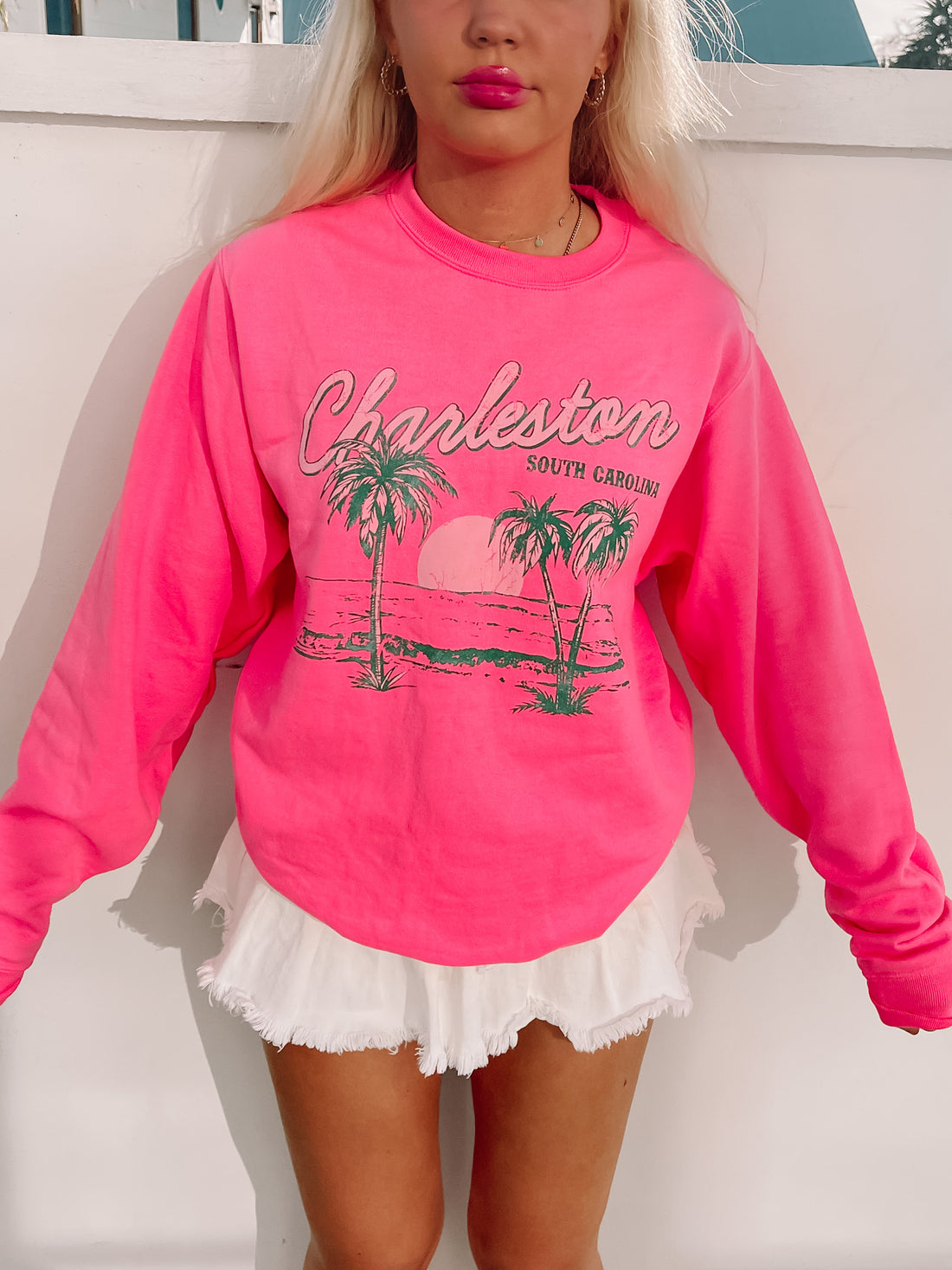 Bright pink crewneck sweatshirt with a palm tree and sunset graphic, styled with a white mini skirt.
