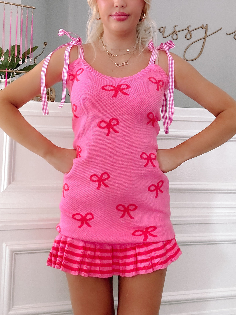 Hot pink knit dress with red bow pattern, gingham straps, and pleated hem.

