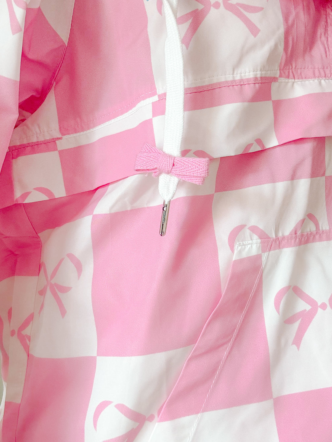 Close-up of pink and white checkered windbreaker's fabric texture; a pink bow adorns the drawstring.
