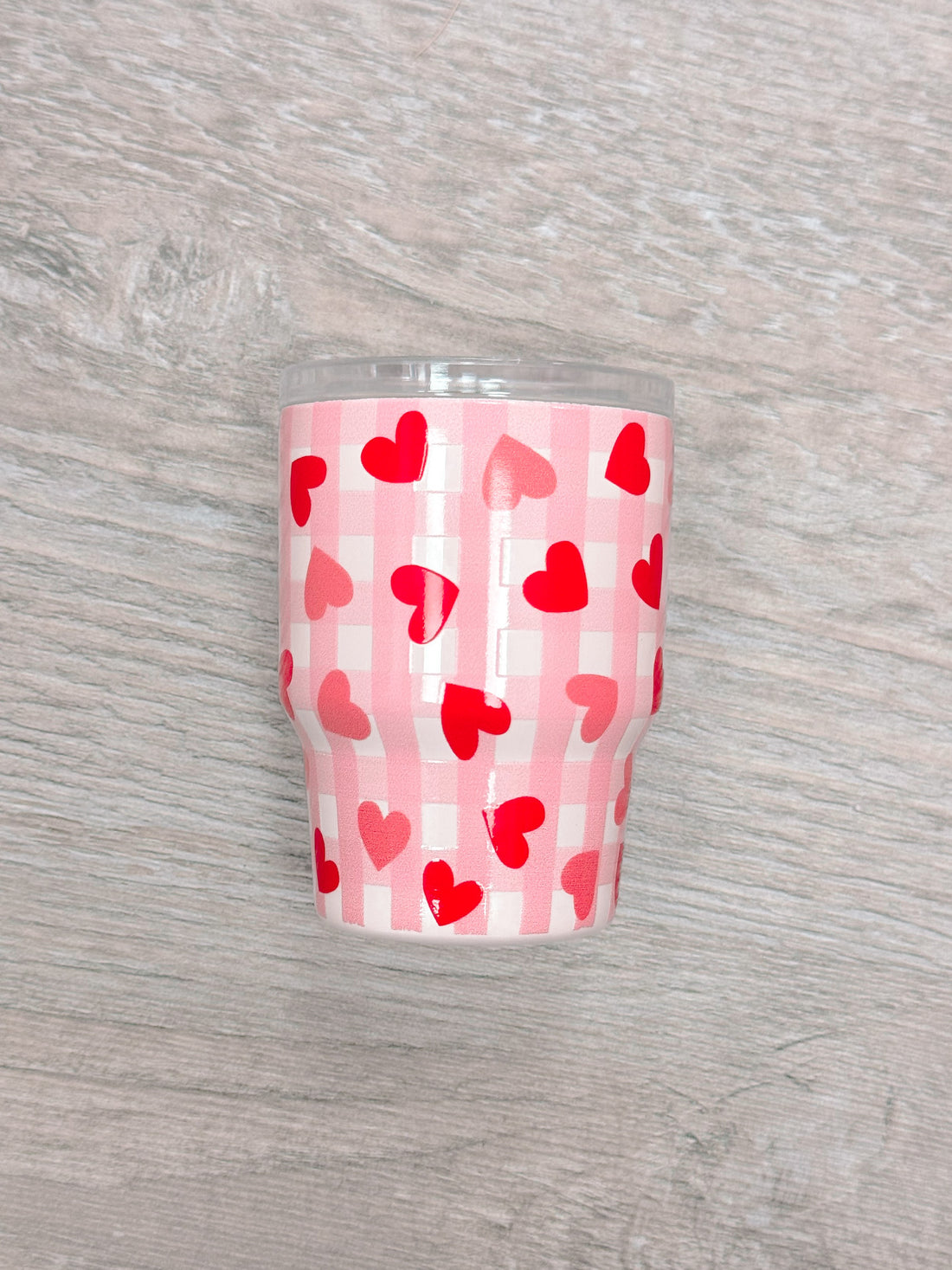 Pink tumbler with red hearts on a gingham pattern, shown on a wood surface.
