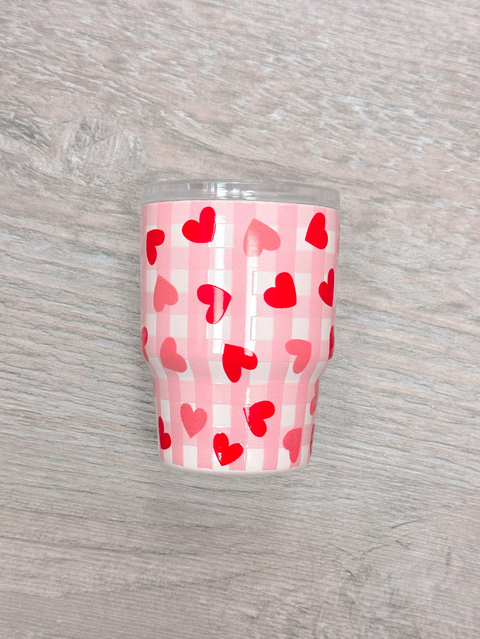 Pink tumbler with red hearts on a gingham pattern, shown on a wood surface.
