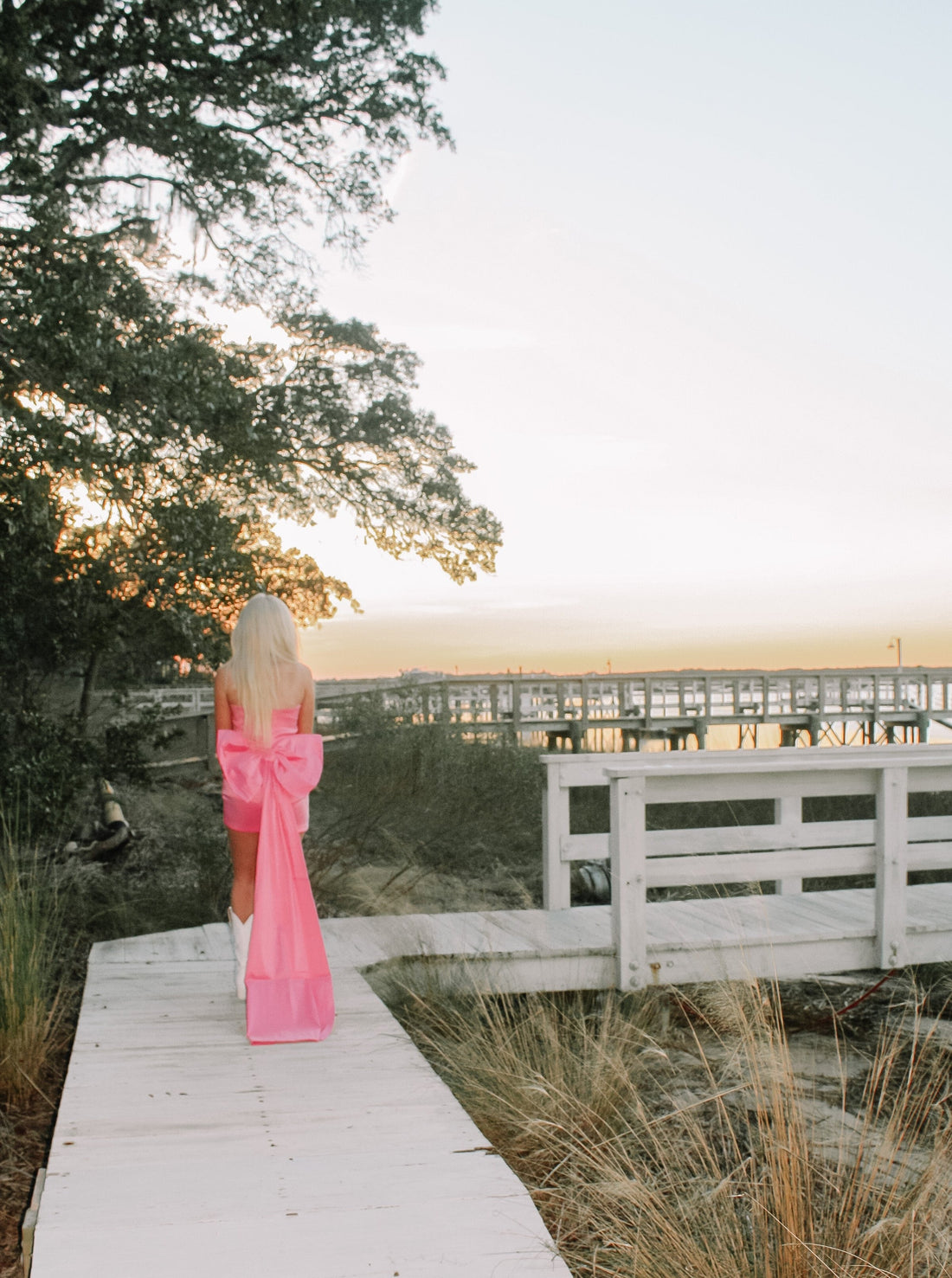 Pink mini dress with large bow detail, worn outdoors near a waterfront boardwalk at sunset.
