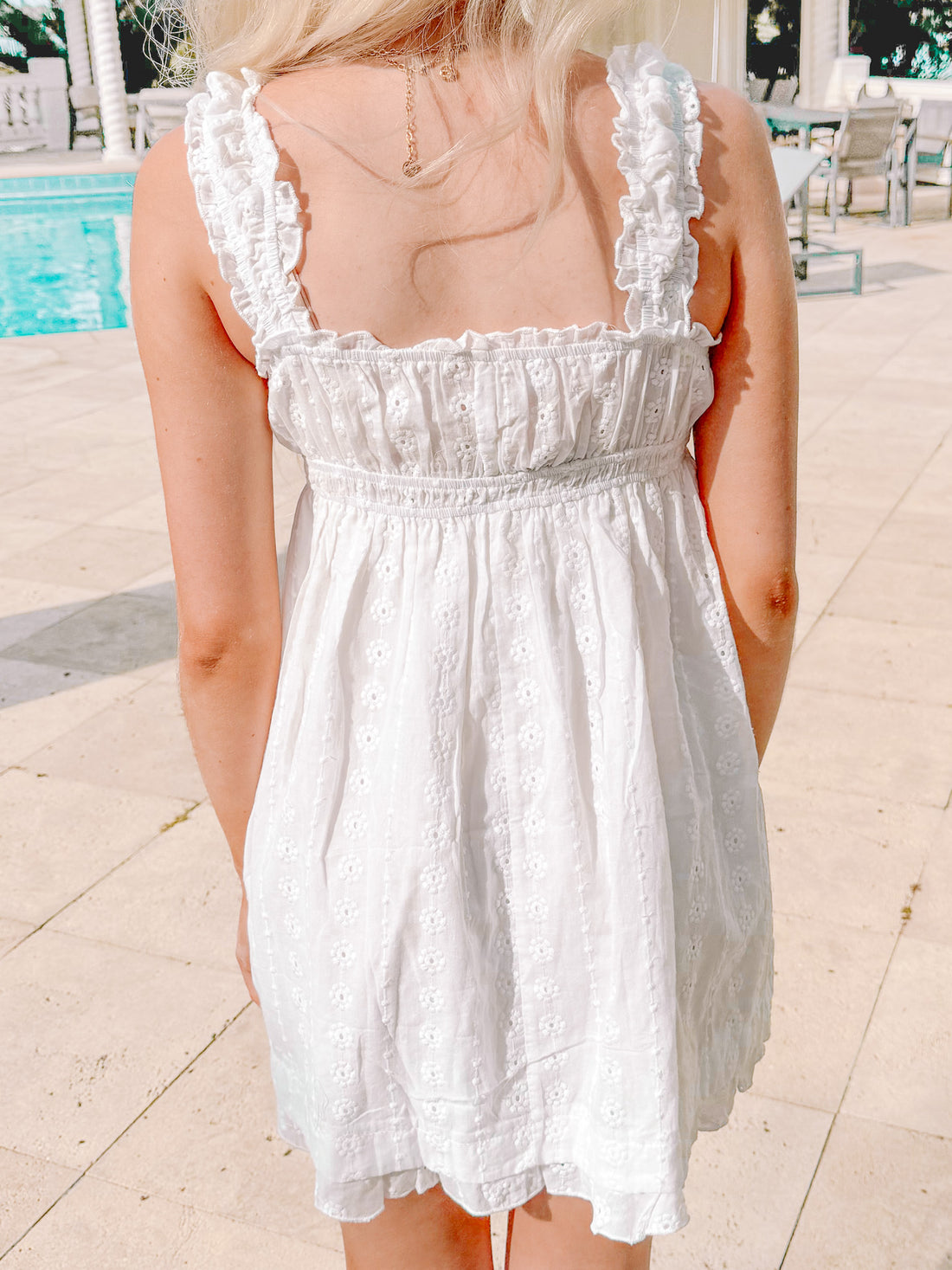 White eyelet mini dress with ruffled straps, shown from the back by a pool.
