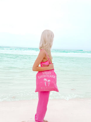 Pink canvas tote bag on a beach, featuring a palm tree design.
