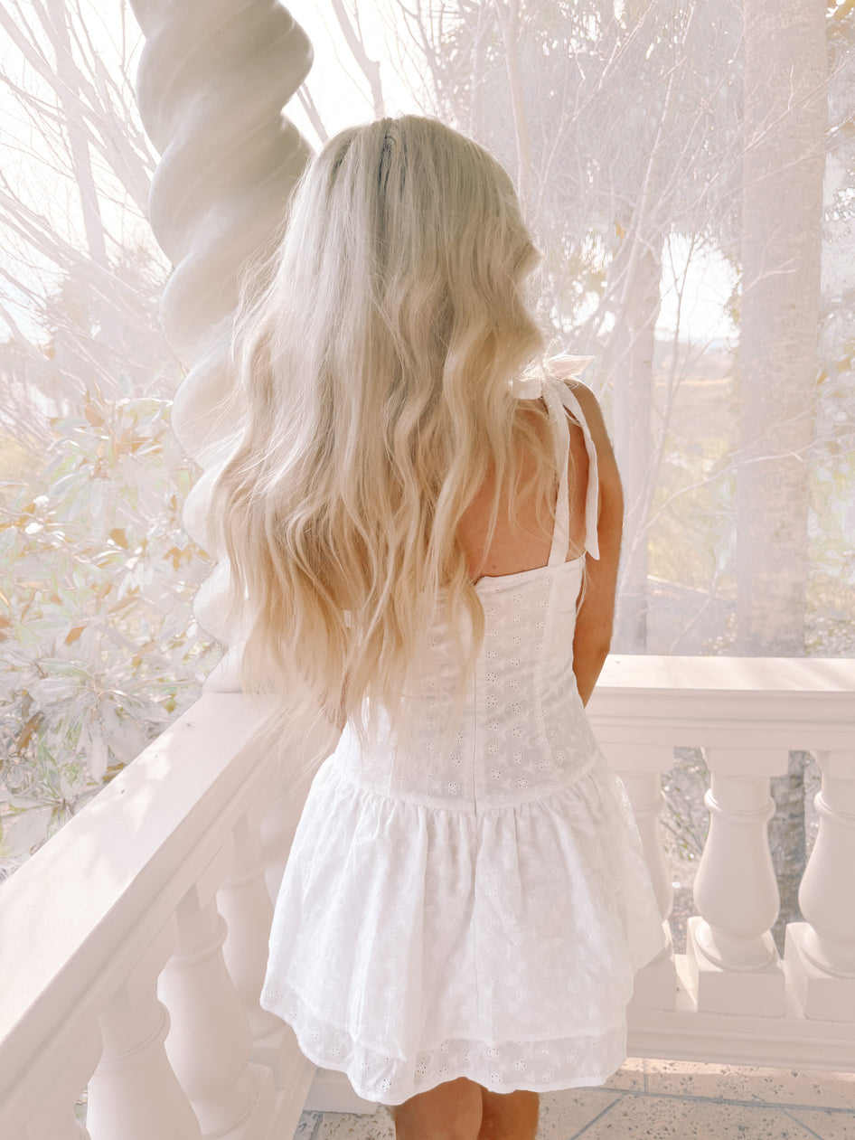 White eyelet mini dress with thin straps, viewed from behind, on a porch overlooking a garden.
