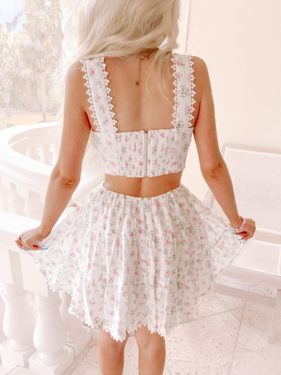 White floral dress with pom-pom trim, shown from behind on a balcony.
