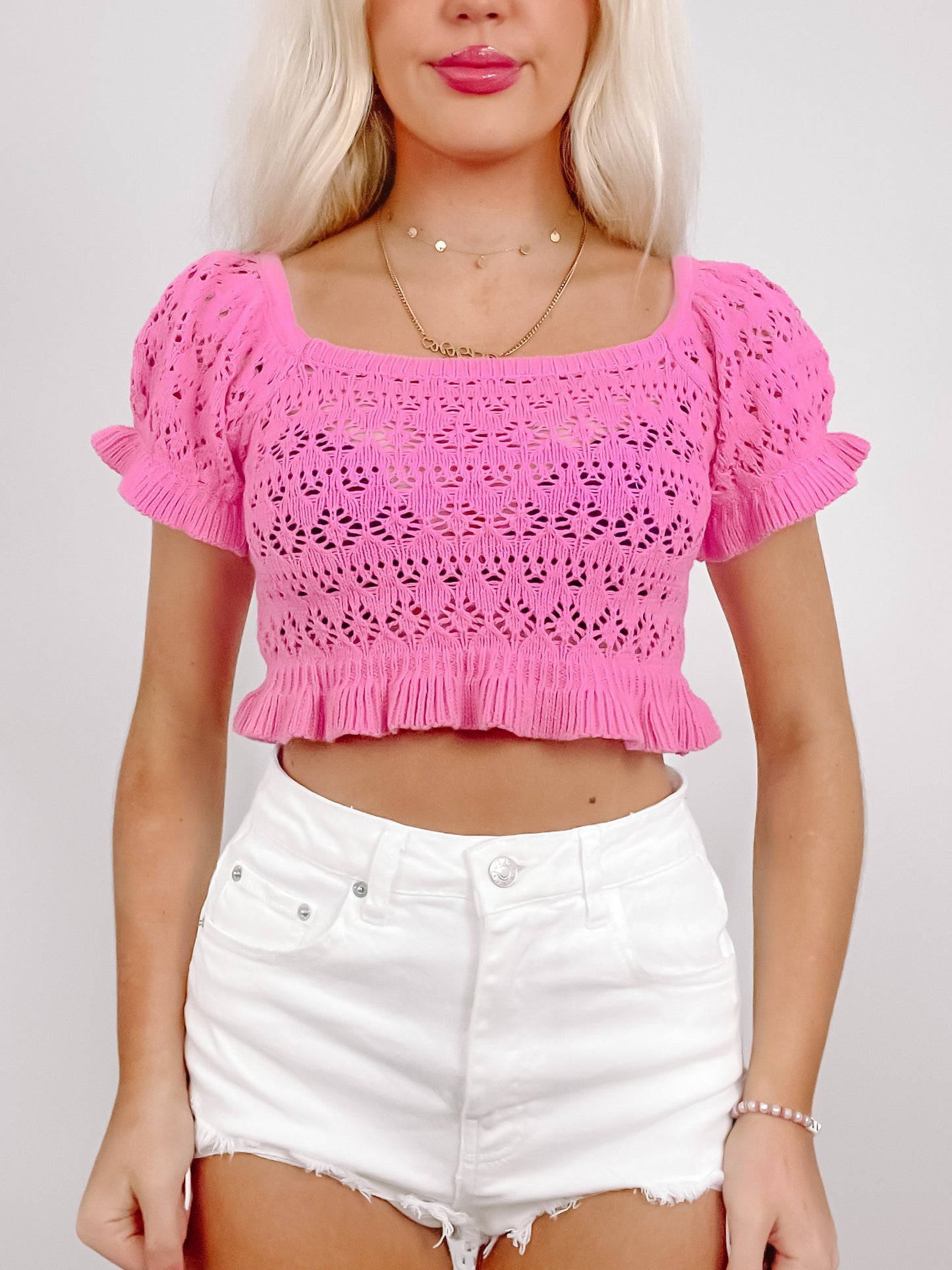 Pink crochet crop top with short sleeves and ruffled hem, modeled with white denim shorts.
