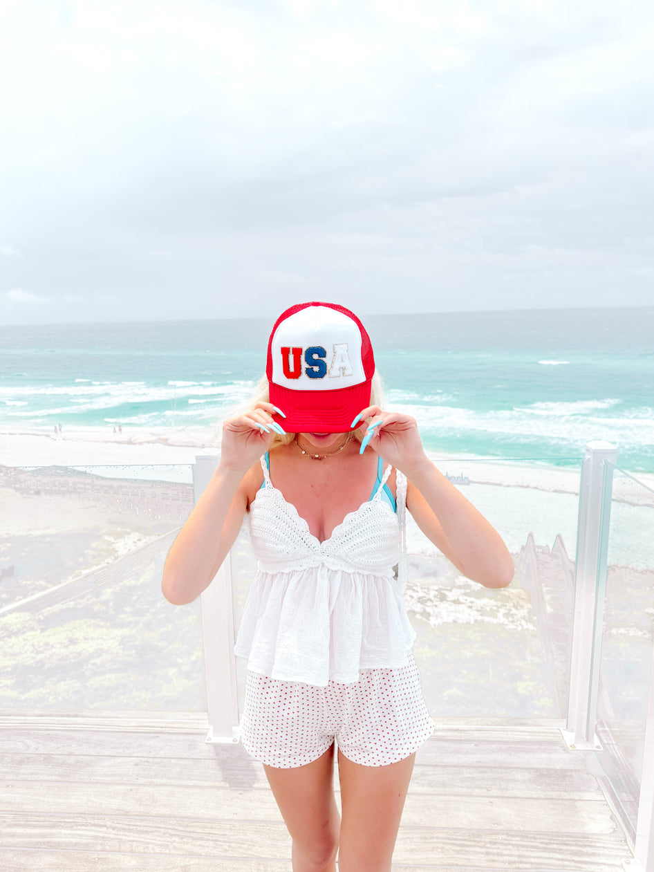Red, white, and blue USA trucker hat worn by a model on a beachside deck.
