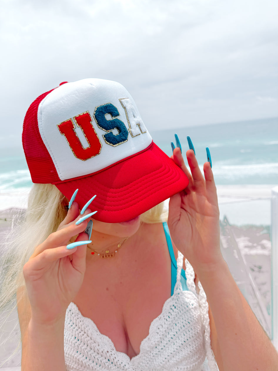 Red, white, and blue USA trucker hat worn by a blonde woman at the beach.
