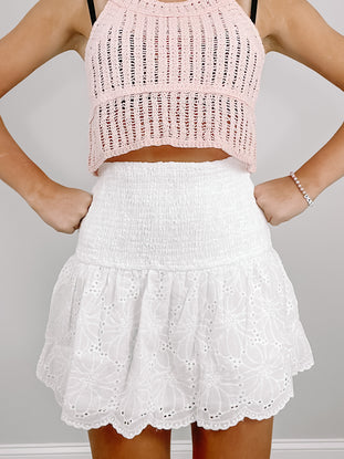 White eyelet mini skirt with smocked waistband, styled with a pink knit top.
