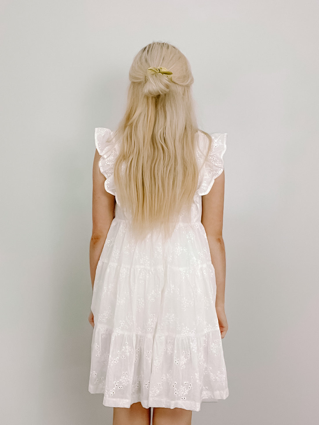 White eyelet mini dress, tiered skirt, ruffle sleeves, shown from the back against a light wall.
