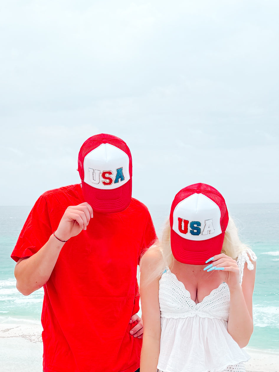 Red, white, and blue "USA" trucker hats worn by a couple at the beach.
