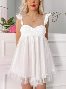 White eyelet mini dress with ruffle trim, showcased against a white paneled wall.
