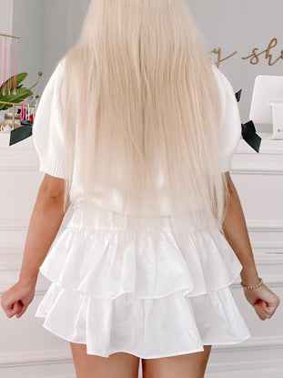 White knit sweater with black bow detail, paired with a layered white ruffle skirt.
