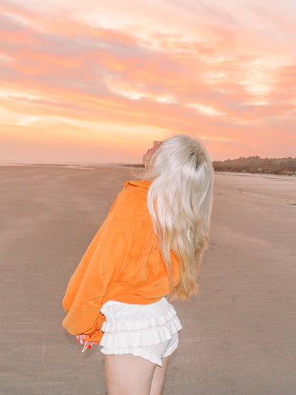 Sunset beach scene featuring a woman in an orange pullover and white ruffled shorts.
