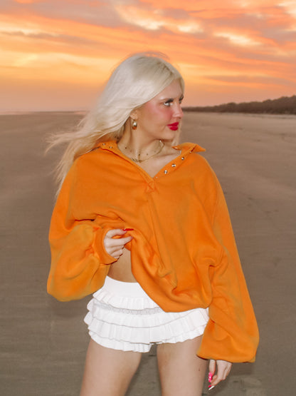 Sunset beach scene featuring a woman in an oversized orange pullover and white ruffled shorts.
