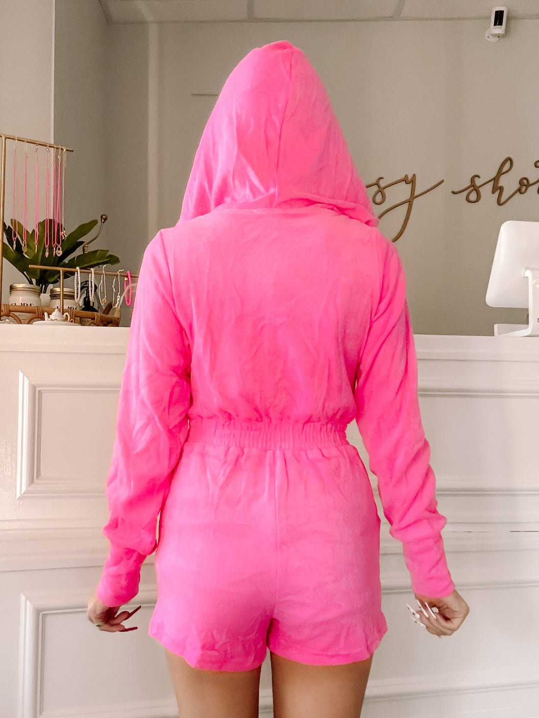 Hot pink hooded jumpsuit, cinched waist, short sleeves, shown from the back in a shop setting.
