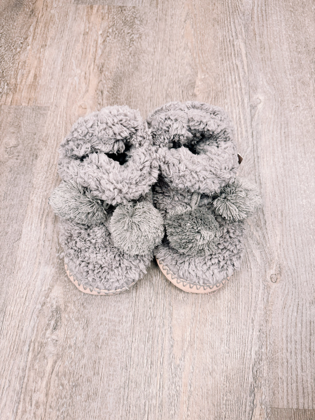 Cozy gray fleece booties with fluffy pom-poms, shown on a wood floor.
