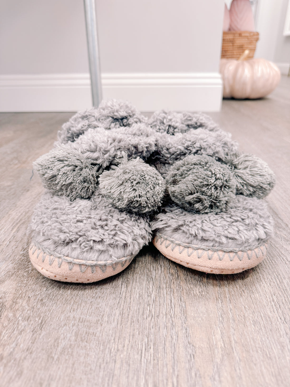 Cozy gray fuzzy slippers with fluffy pom-poms, resting on a wood floor.
