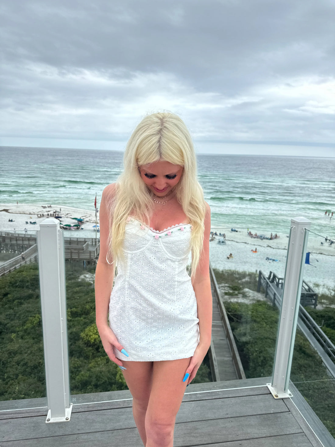 White eyelet mini dress with floral accents, worn outdoors near the ocean.
