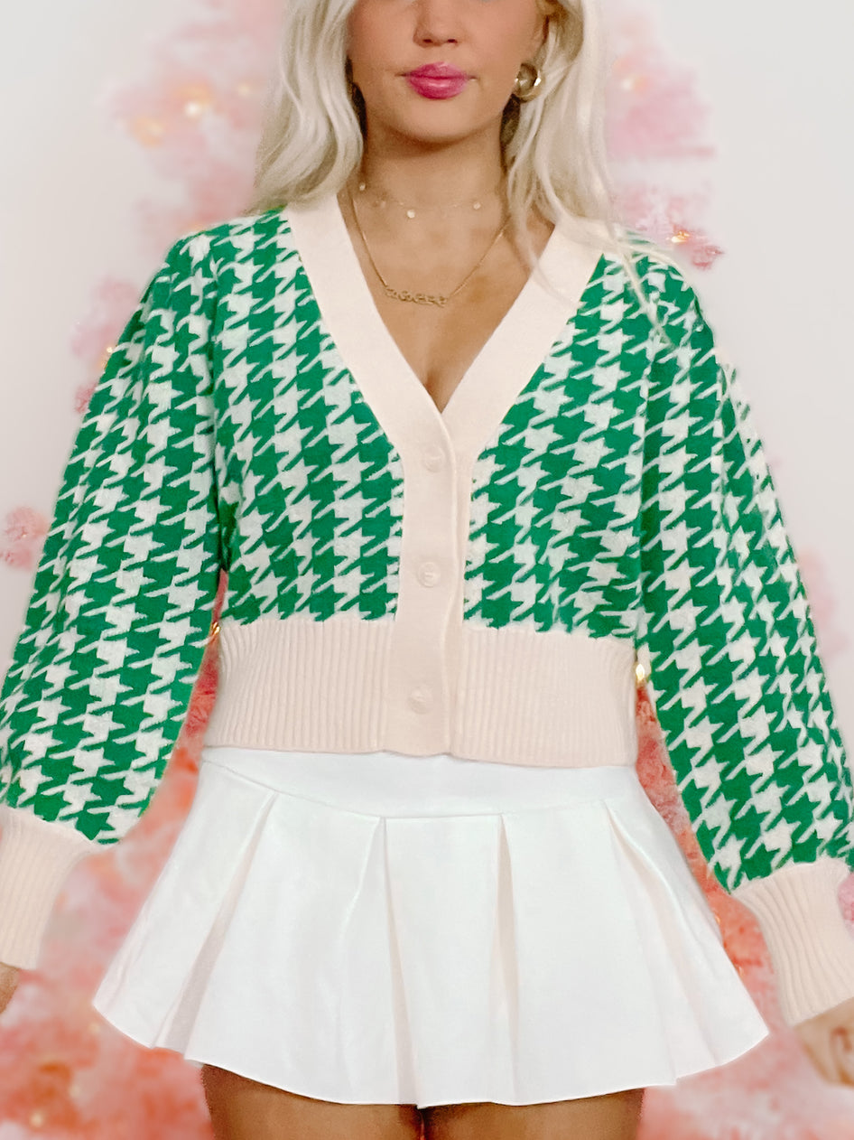 Green and white houndstooth cardigan with a cream-colored bottom band, shown on a model against a pink background.
