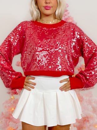 Red sequin sweater, festive and sparkly, shown on model with white pleated skirt.
