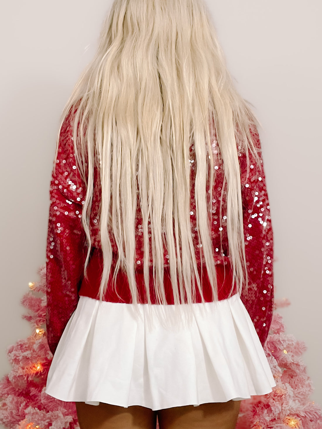 Red sequin sweater with white pleated mini skirt, shown from the back against a pink Christmas tree.
