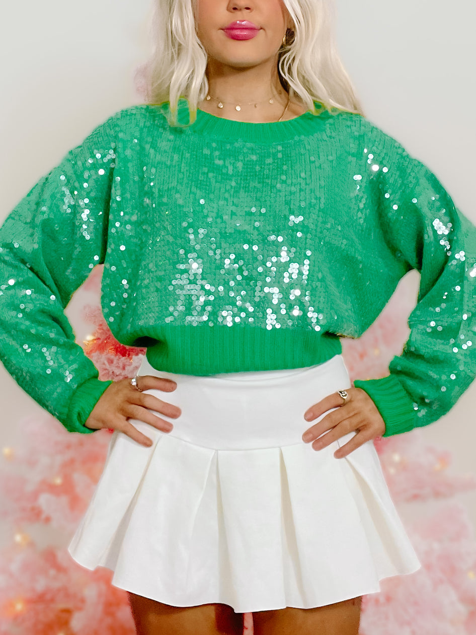 Green sequin sweater with long sleeves, modeled against a pink backdrop.
