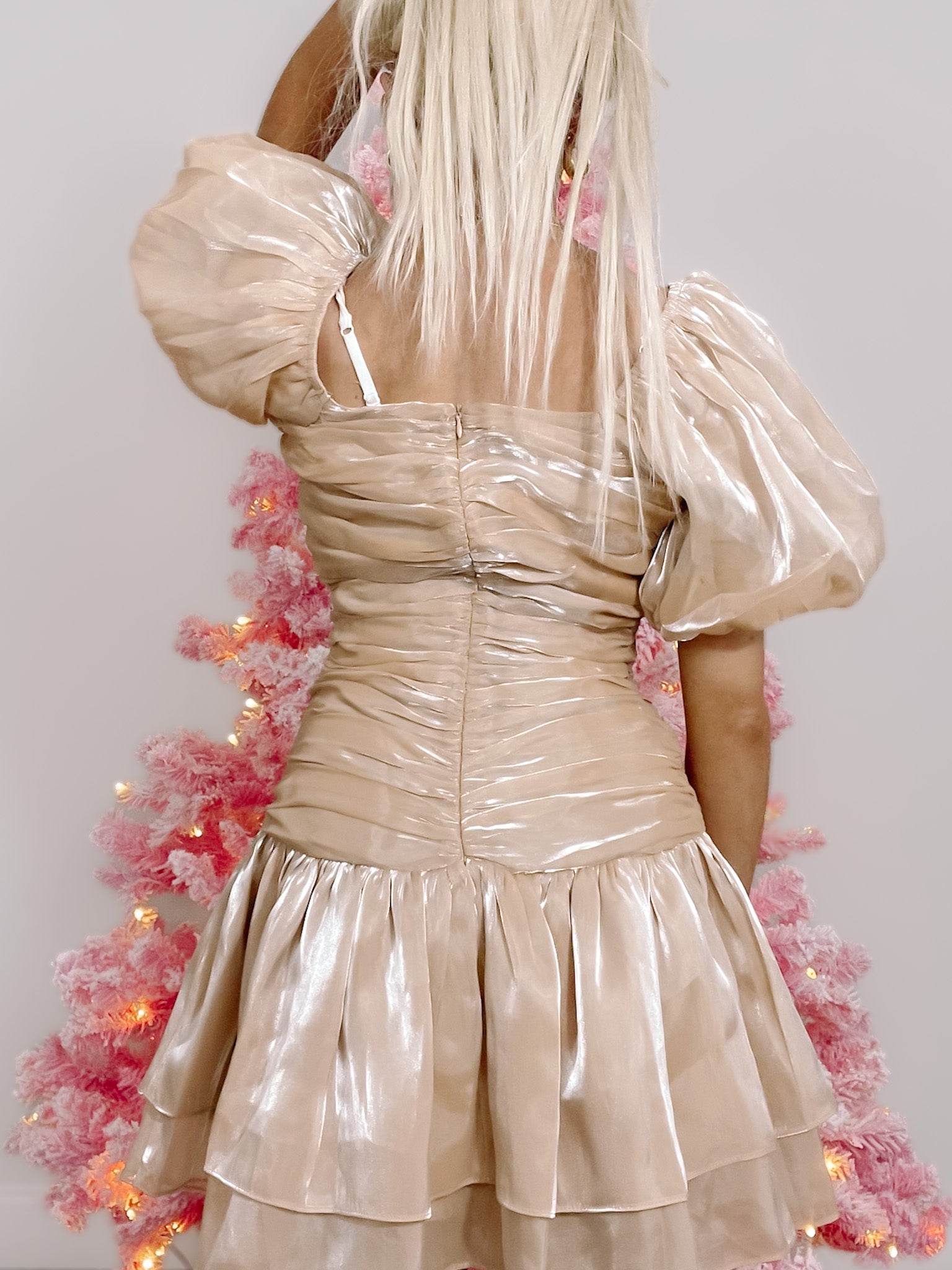 Champagne-colored mini dress with puff sleeves, tiered skirt, and a fitted bodice, shown against a pink Christmas tree.
