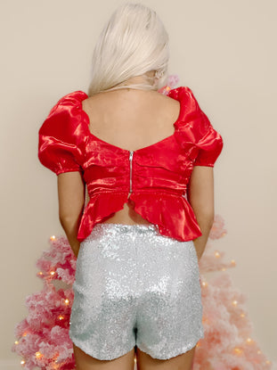 Red satin puff-sleeve crop top paired with sparkly silver shorts, shown from the back against a pink Christmas tree.
