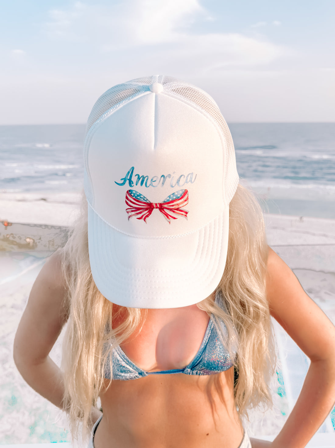 White trucker hat with patriotic bow and "America" lettering, ocean backdrop
