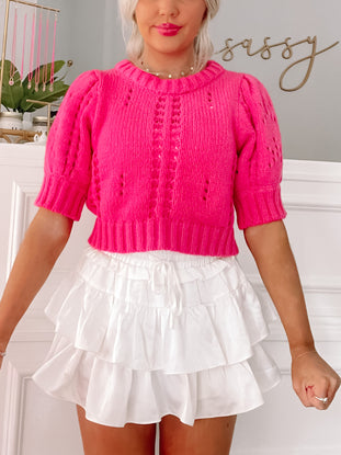Hot pink cropped knit sweater with eyelet detailing, paired with a white tiered ruffle mini skirt.
