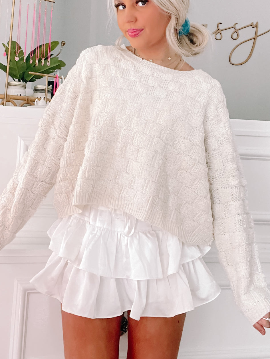 Cream cable-knit sweater paired with white ruffle shorts; lifestyle shot.
