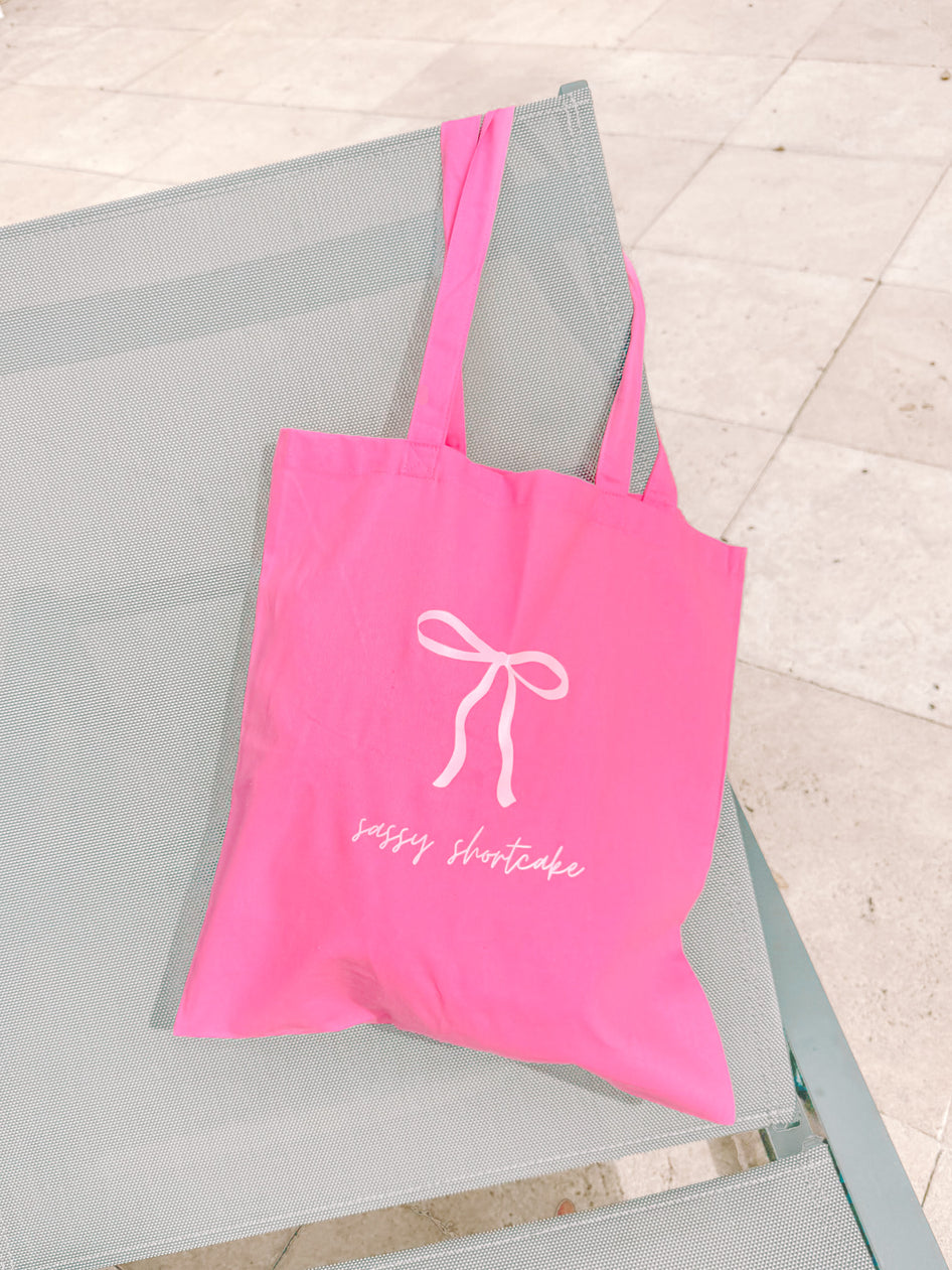 Pink canvas tote bag with bow design, resting on a gray chair outdoors.

