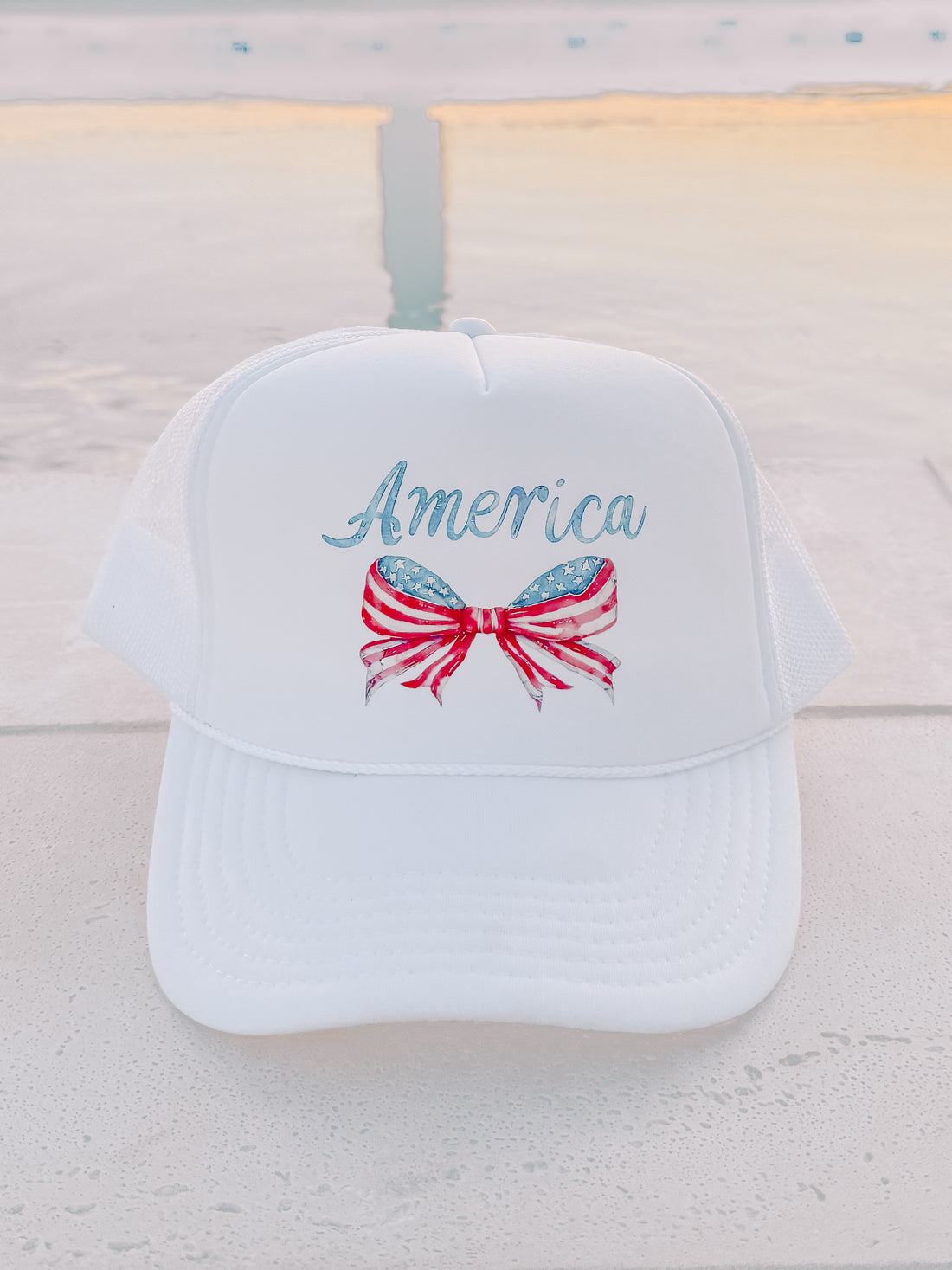 White trucker hat with patriotic bow and "America" design, shown outdoors near water.
