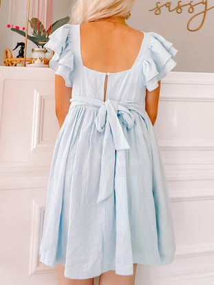 Light blue mini dress with ruffled sleeves and a tie waist, shown from the back against a white wall.
